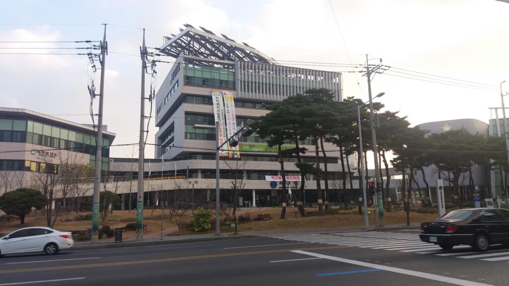 a white car driving down a street with a building in the background - File:Gwangju Seo-gu Office.jpg