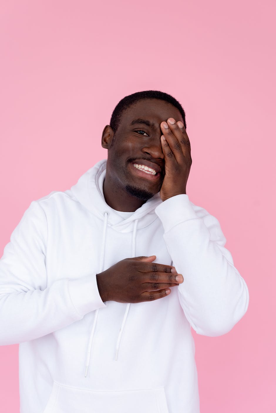Cheerful man wearing white hoodie covering face with hand and looking at the camera